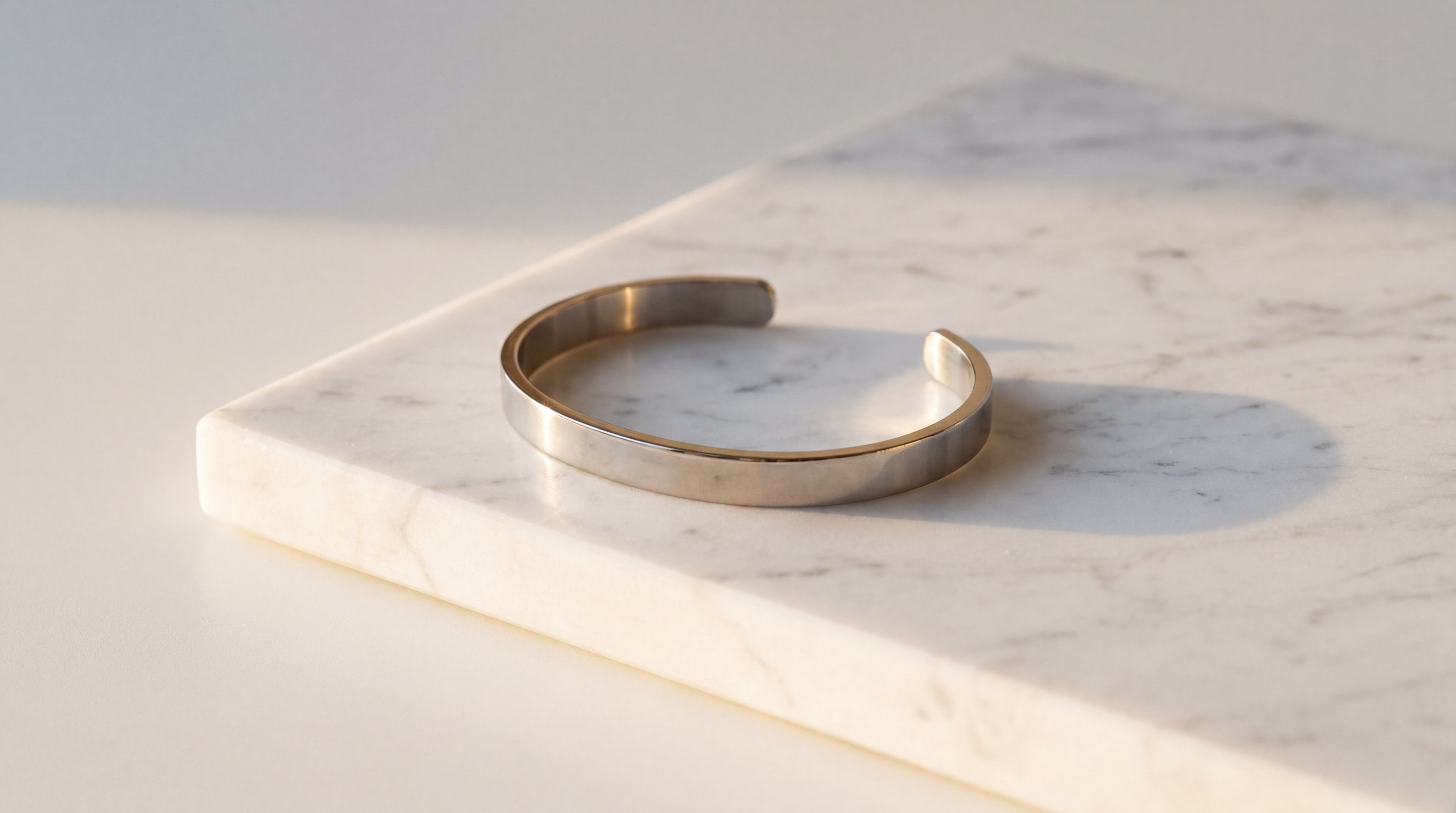 Bracelet manchette en acier inoxydable 316L posé sur une surface en marbre blanc, mise en valeur artistique et lumière rasante dorée, rendu luxueux et minimaliste