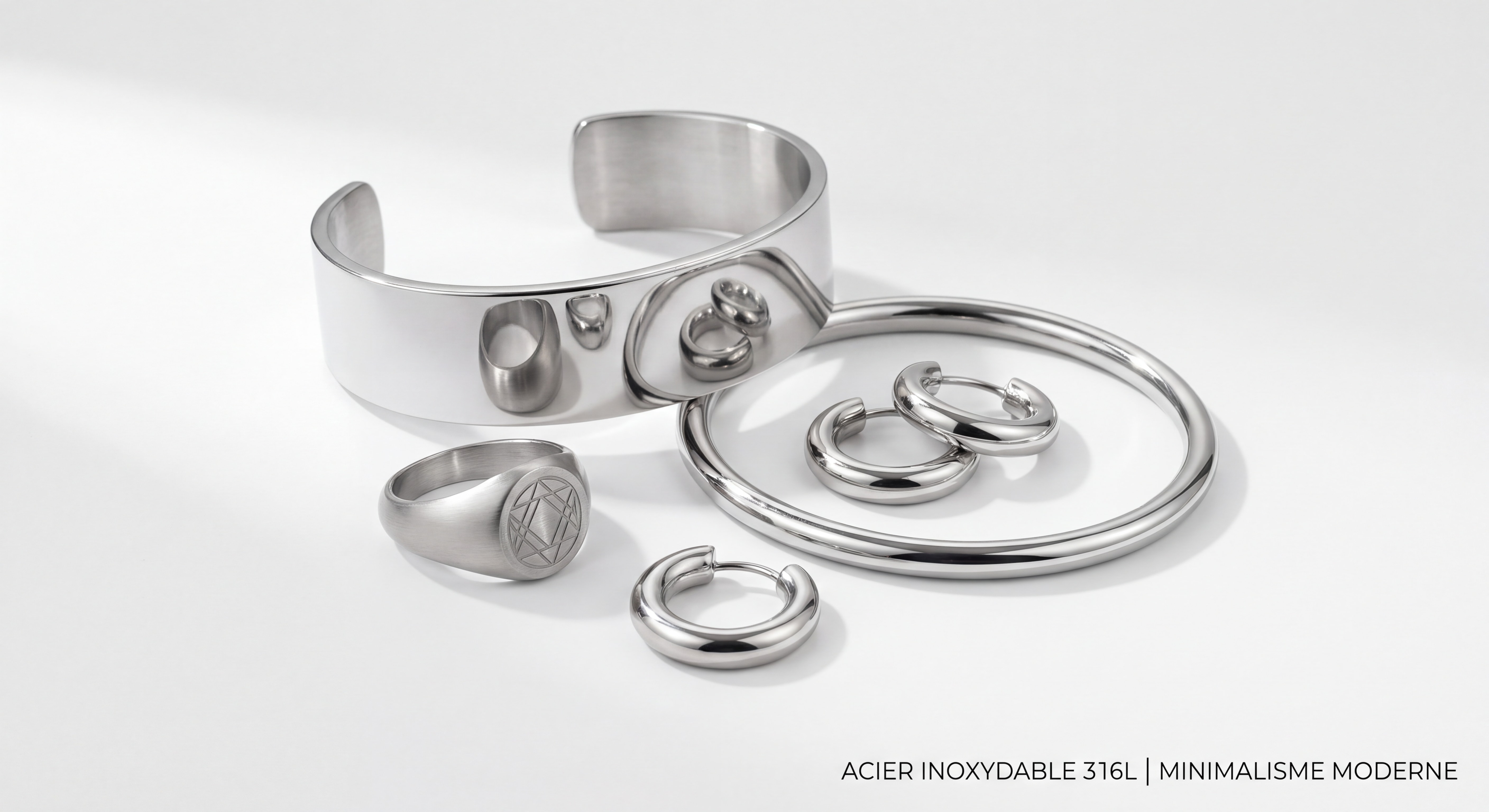 Plusieurs bijoux en acier inoxydable 316L — bracelet manchette, chevalière, créoles et jonc — disposés élégamment sur fond blanc épuré, style éditorial minimaliste