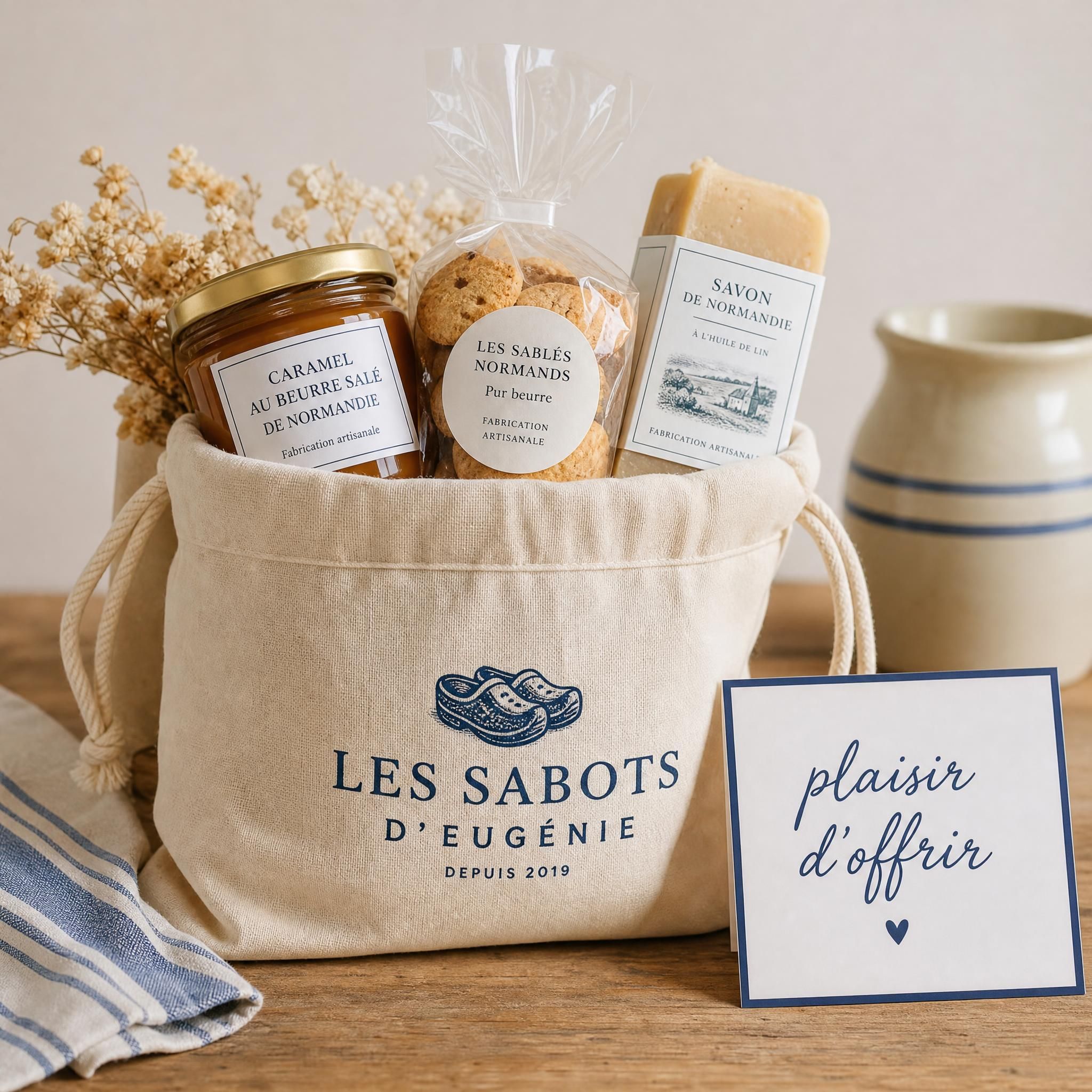 Pochon textile Les Sabots d'Eugénie rempli de produits du terroir normand — caramel au beurre salé, biscuits artisanaux, savon normand — posé sur une table en bois avec une carte